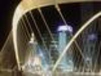 Foto: Blick bei Nacht von einer Hängebrücke auf die Halteseile. Im Hintergrund beleuchtete Hochhäuser.