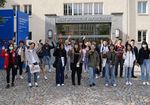 Eine Gruppe internationaler Studierende steht vor Hochschuleingang und winkt in die Kamera.
