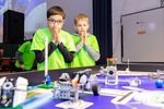 Zwei Schüler stehen mit gefalteten Händen hinter einem Lego-Spielbrett auf dem Roboter bestimmte Aufgaben erfüllen müssen.