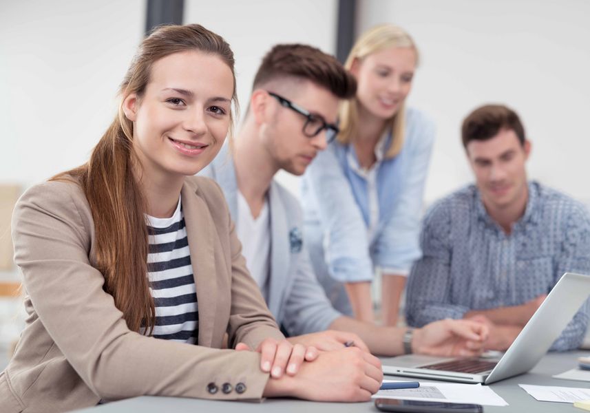 Eine Frau bilckt in die Kamera. Hinter ihr eine Arbeitsgruppe an einem Laptop. 