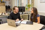 Foto: Junger Mann, junge Frau, sitzen an einem Tisch in einem Büro und sprechen, vor ihnen eine Bewerbungsmappe und ein Laptop.
