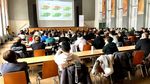 Gut gefüllte Aula. Die Studierenden sitzen an Tischen und verfolgen die Präsentation auf einer Leinwand.