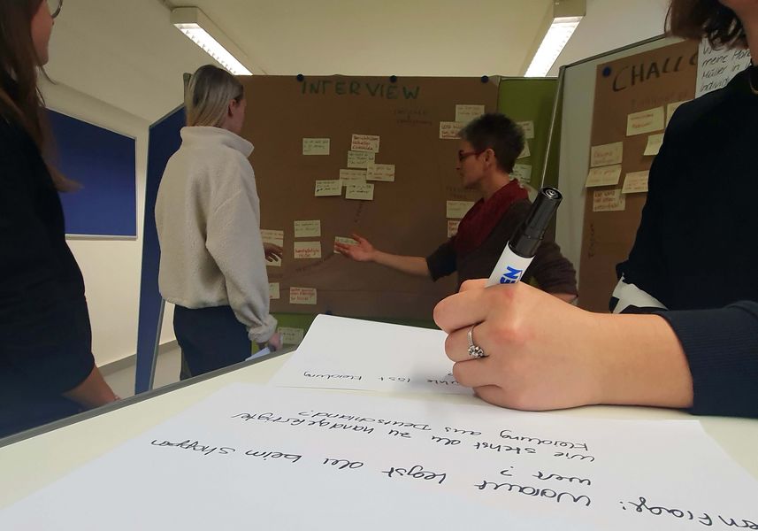 Workshop-Situation. Person schreibt mit Stift auf ein Papier, im Hintergrund ein Flipchart