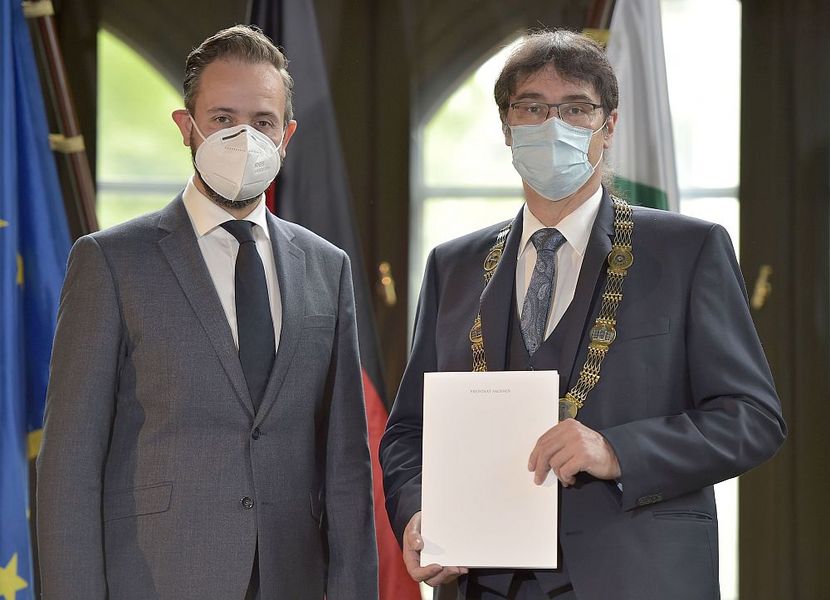 Foto: Zwei Männer mit Maske schauen in die Kamera. Der Mann auf der rechten Seite hält ein Papier vor sich. 