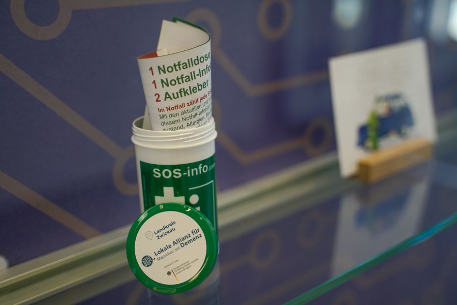 Auf dem Foto ist die Notfalldose abgebildet. Im Hintergrund erkennt man eine Postkarte zur Thematik Demenz.