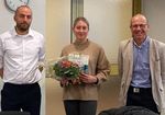 3 Personen lächeln in die Kamera, Person Mitte hat Blumenstrauß und Urkunde in der Hand.