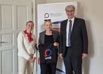 3 Personen stehen vor einem hellen Hintergrund und sind fröhlich. Die Person in der Mitte, eine Frau hält einen Beutel mit Hochschullogo hoch.