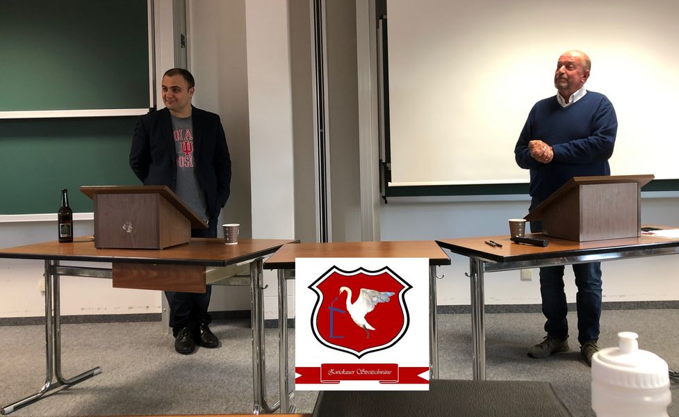 Foto: Showdebatte zwischen Hr. Prof. Kolev und Hr. Sullivan. Beide stehen in einem Raum vor einem Tisch mit Rednerpult Aufsatz.