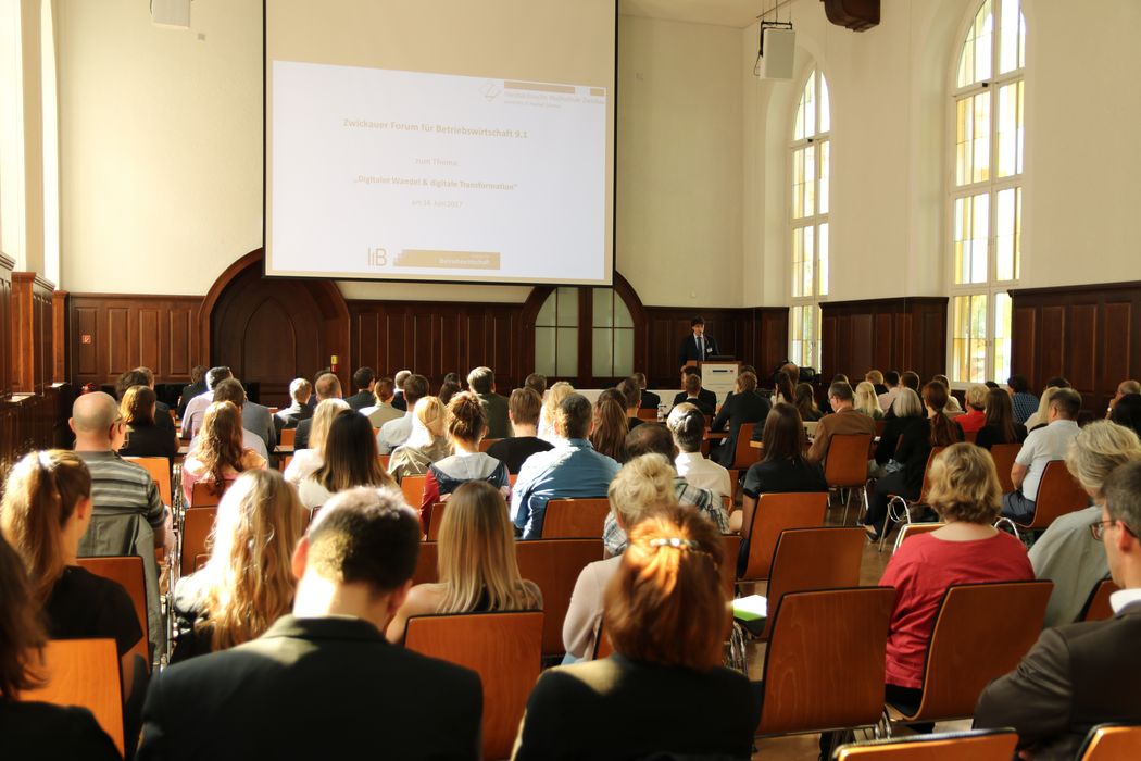 Foto: Teilnehmer sitzen in der Aula und hören einem Vortrag zu.
