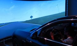 Foto: Fahrsimulator. Eine Schülerin sitzt in einem PKW. Auf einer Leinwand vor ihr eine Straße projiziert, welche Sie gerade befährt.