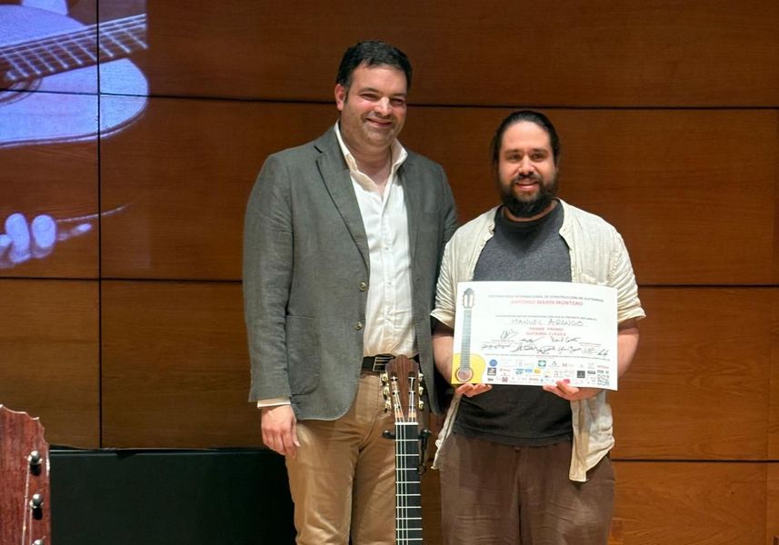 Zwei Personen stehen auf einer Bühne und blicken in die Kamera. Eine Person hält eine Urkunde in der Hand. Im Vordergrund steht eine Gitarre.