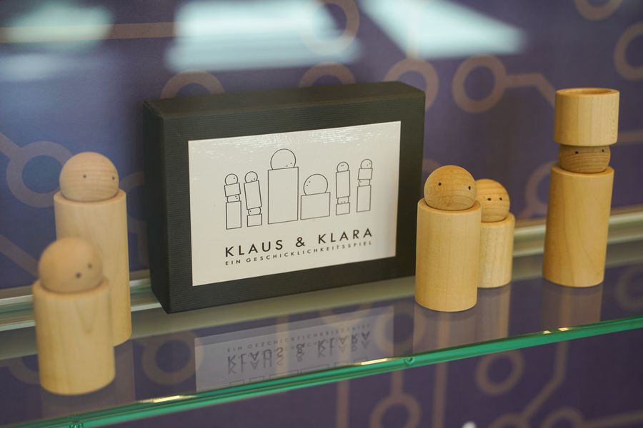 Auf dem Foto sieht man die Holzfiguren zum Stapeln auf einem Glasregal stehen. Es sind Holzzylinder als Körper und Holzkugeln als Kopf. Die schwarze Box steht dabei.