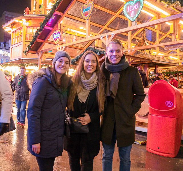 Gruppenfoto: Drei Studierende vor einer Hütte auf dem Weihnachtsmarkt.