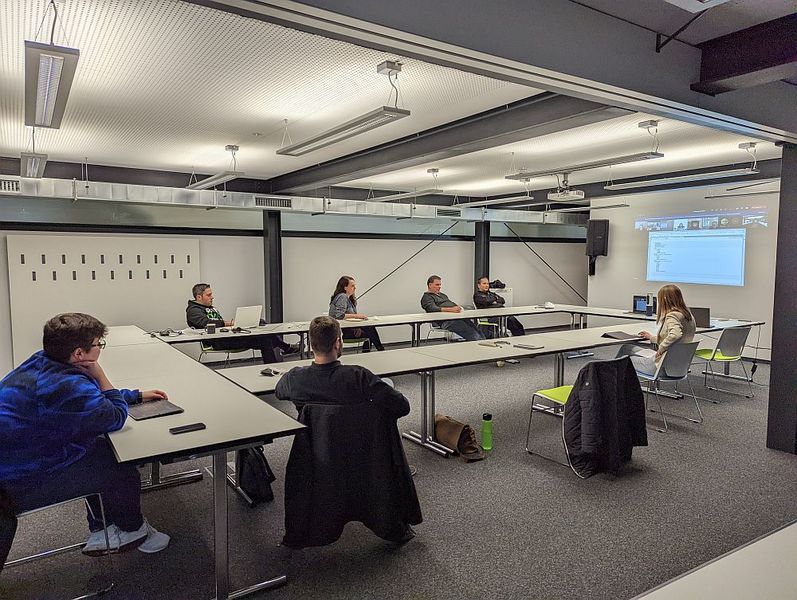 Foto: In einem großen Beratungsraum stzen einige Menschen weit auseinander. Auf einer Leinwand sind weitere Personen zugschalten. 