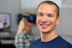 Foto: Prof. Sven Hellbach blickt lächelnd in die Kamera. Er trägt ein blaues T-Shirt. Im Hintergrund ist verschwommen eine Studierende mit einer VR-Brille zu sehen.