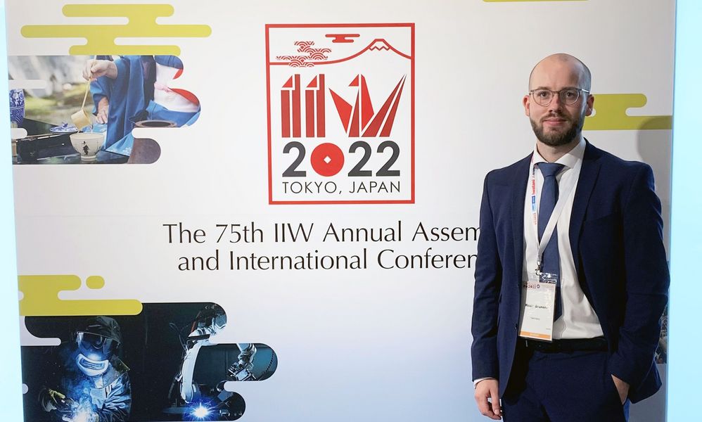 Ein Mann im Anzug steht vür einem Logo der "IIW Annual Assembly and International Conference on Welding and Joining", Tokyo 2022