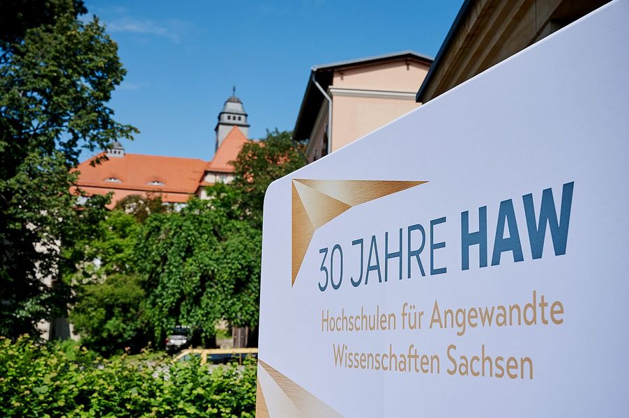 Ein Schild mit der Ausfschrift 30 Jahre Hochschulen für Angewandte Wissenschaften steht vor einem Gebäude, das teilweise von Bäumen und Sträuchern verdeckt wird.