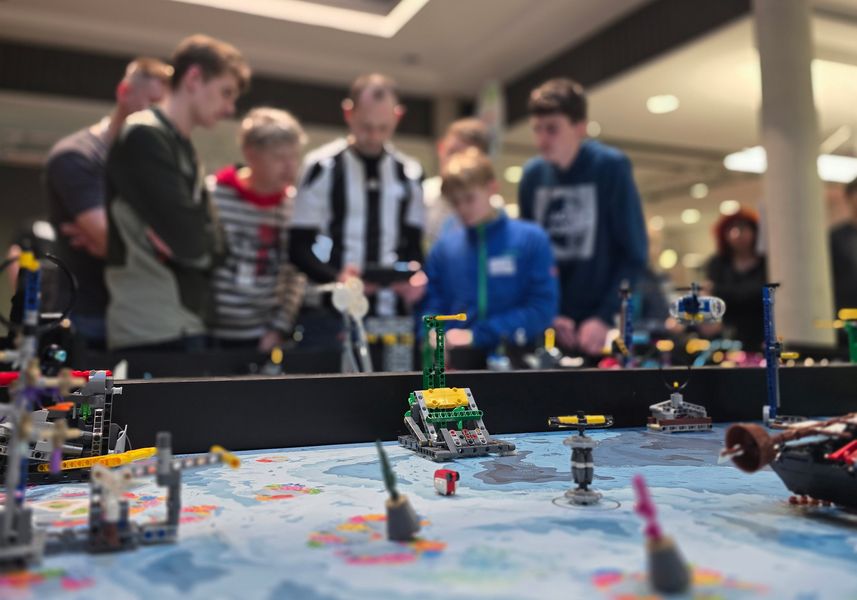 Personen stehen hinter einem Lego-Spieltisch, auf dem sich Roboter bewegen und Aufgaben lösen. 