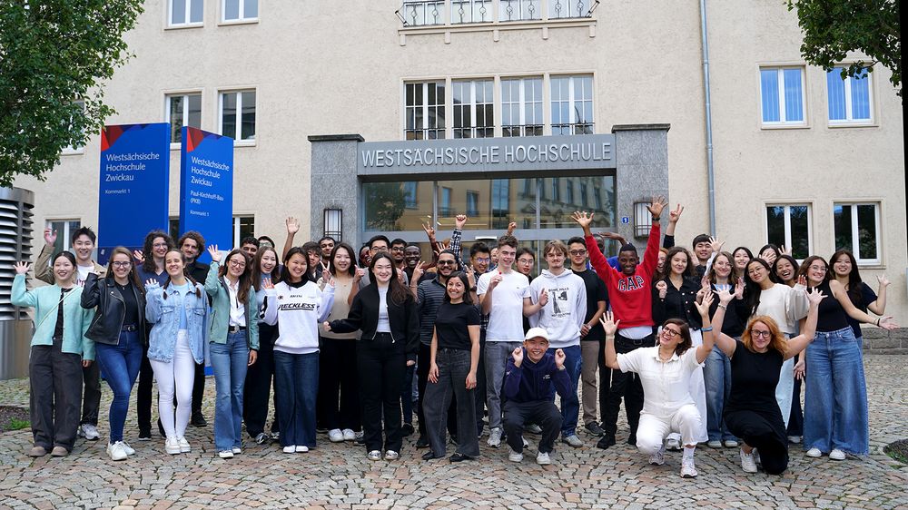 Eine Gruppe internationaler Studierender steht vor dem Eingang der Hochshcule und winkt in die Kamera. 