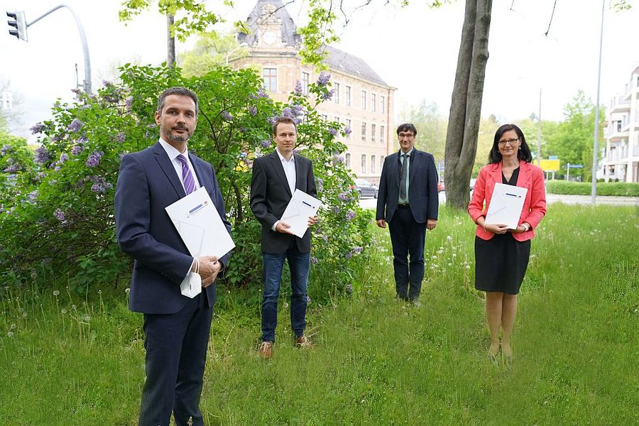Foto: Auf einer Wiese stehen vor einem Strauch links zwei Männer mit Urkundenmappen in ihren Händen, rechts daneben Rektor Prof. Kassel und ganz rechts eine Frau mit einer Urkundenmappe in den Händen.