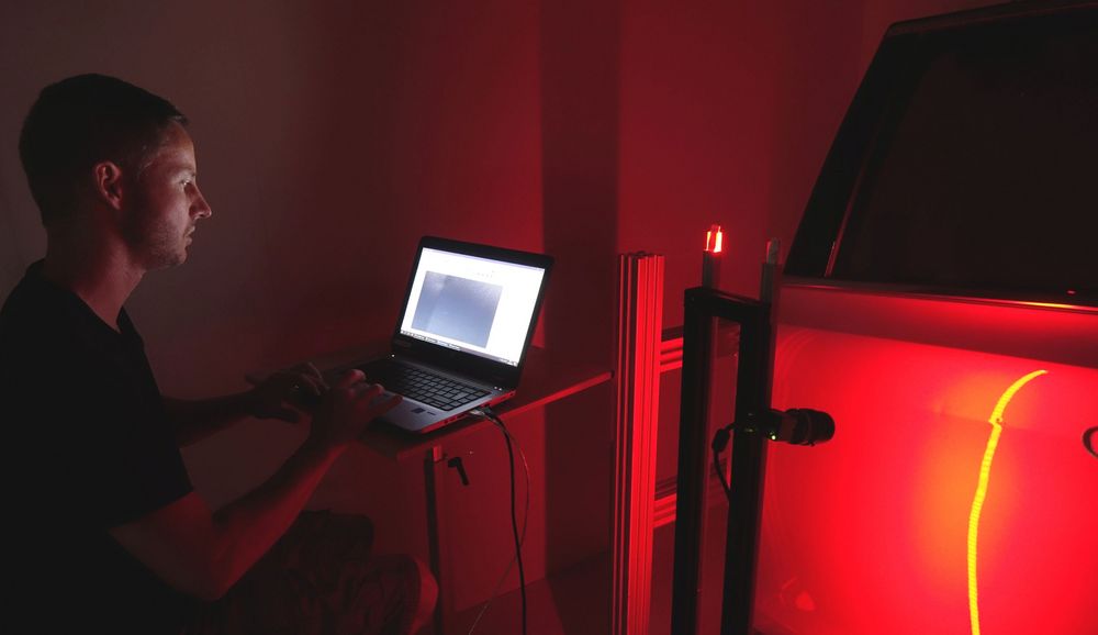Foto: Ein Mann sitzt im Dunkeln vor einem Laptop. Daneben ist ein in rot getauchtes Fahrzeug zu sehen. 