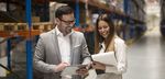 Mann und Frau im Business-Look stehen in einem Hochregallager und unterhalten sich. Er hält ein Tablet in der Hand, sie einen Notizblock.