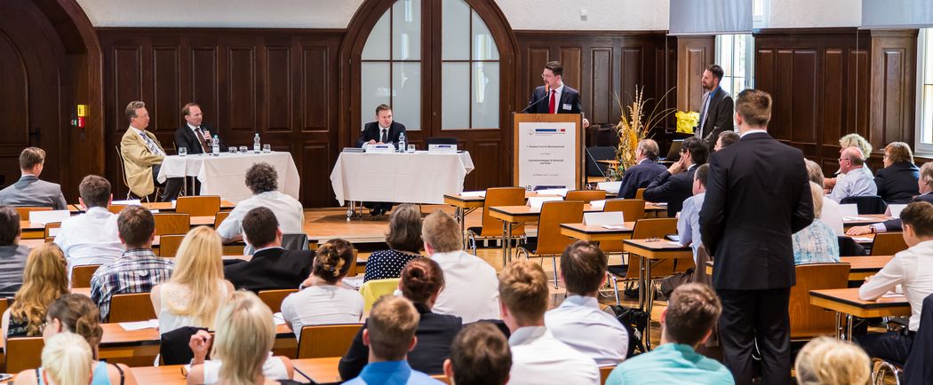 Foto: Teilnehmer sitzen in der Aula und hören einer Podiumsdiskussion zu.
