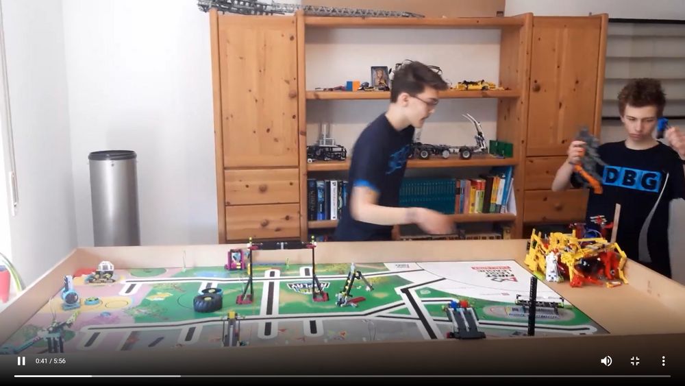 Foto: Im Vordergrund steht eine Lego-Fläche, im Hintergrund bewegen sich zwei Jungen während des Robot Games.