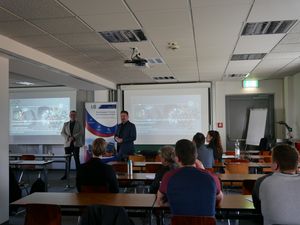 Foto: Studierende an Tischen hören zwei Personen zu