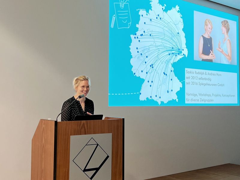Eine Frau steht hineter einem Pult an einem Mikrofon und spricht mit einem Lächeln im Gesicht. 
