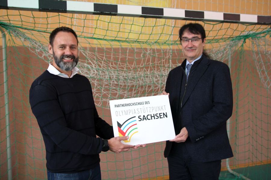 Foto: Zwei Männer (einer Rektor Prof. Kassel) halten vor einem Tor gemeinsam ein Schild mit der Aufschrift Partnerhochschule des Olympiastützpunktes Sachsen e.V.