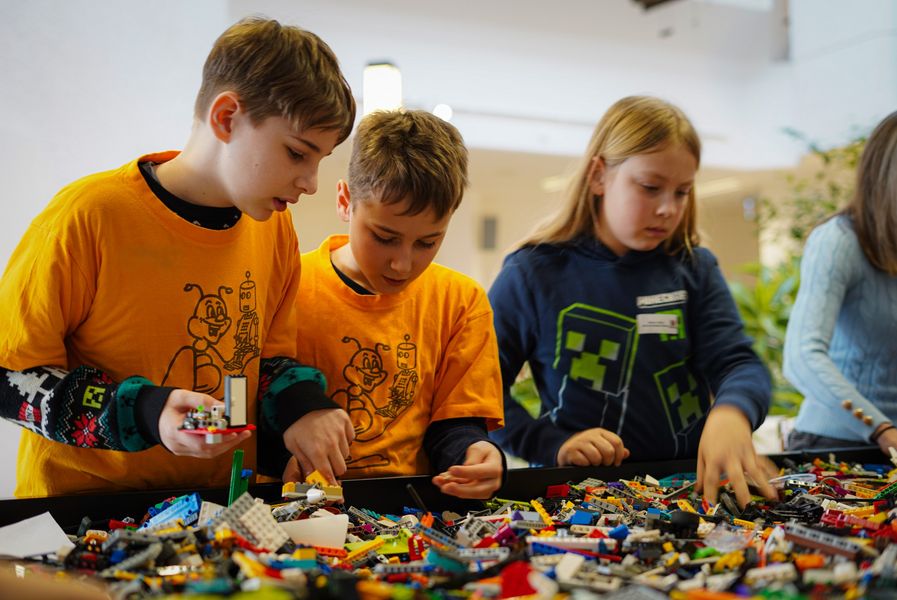 3 Kinder wühlen suchend auf einem Tisch. Auf diesem liegen verschiedene Lego Bausteine vertreut. 