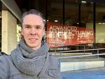 Christopher Taudt steht vor einem Bürogebäude. Im Schaufenster ist ein Transparent mit der Aufschrift "Advanced Manufacturing Precinct" angebracht.