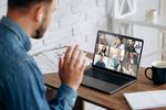 Foto: Ein Mann winkt vor einem Laptop, auf dem Bildschirm sind mehrere Menschen in einer Videokonferenz und winken ebenfalls.