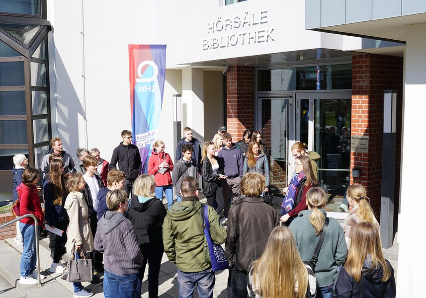Personen warten vor einem Eingang mit der Aufschrift Hörsäle, Bibliothek. 