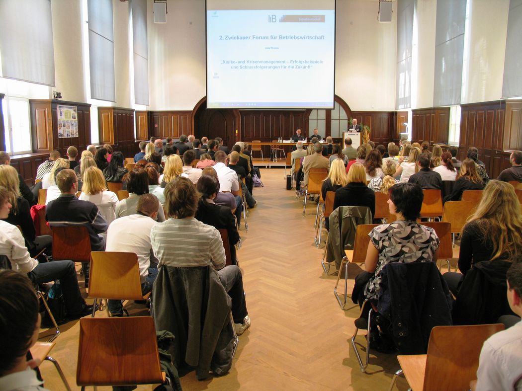 Foto: Teilnehmer sitzen in der Aula und hören einem Vortrag zu.