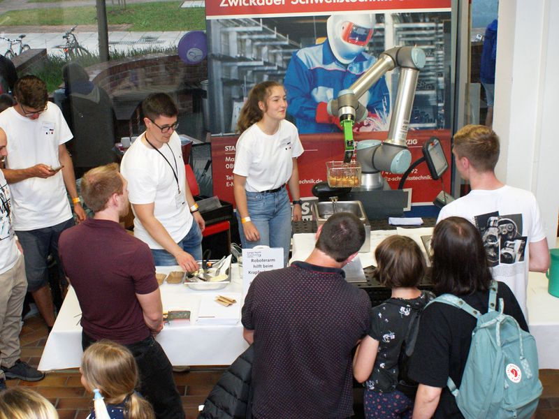 Studenten der Hochschule im weißen T-Shirt führen Roboterarm vor, der Krapfen aus der Fritteuse holt