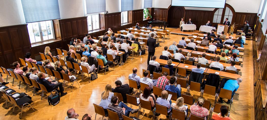 Foto: Teilnehmer sitzen in der Aula und hören einer Podiumsdiskussion zu.