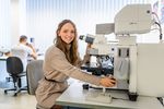Mädchen mit langen Haaren sitzt vor einem Mikroskop und schaut in die Kamera