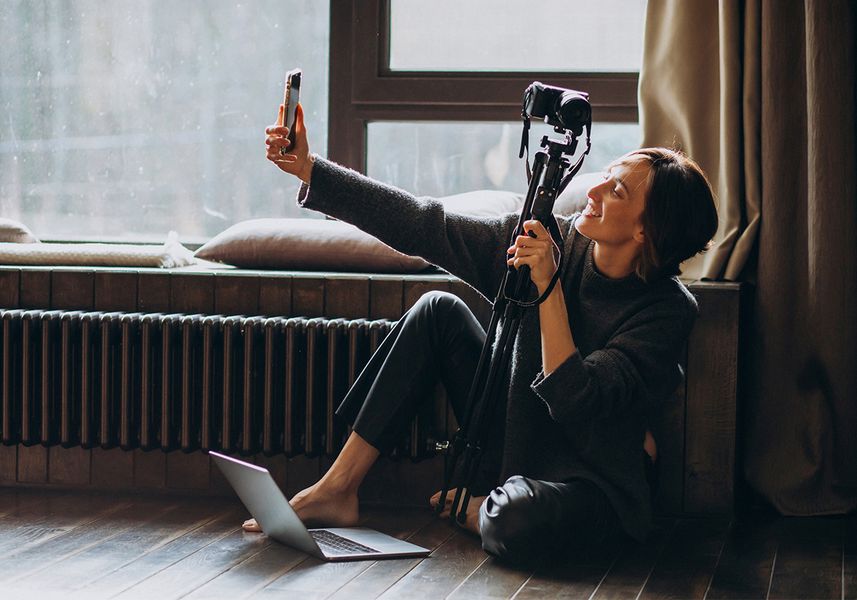 Eine Studierende macht ein Selfie mit einem Smartphone in ihrer Wohnung. In der anderen Hand hät sie eine Digitalkamera. Zu ihren Füßen steht ein Laptop. 