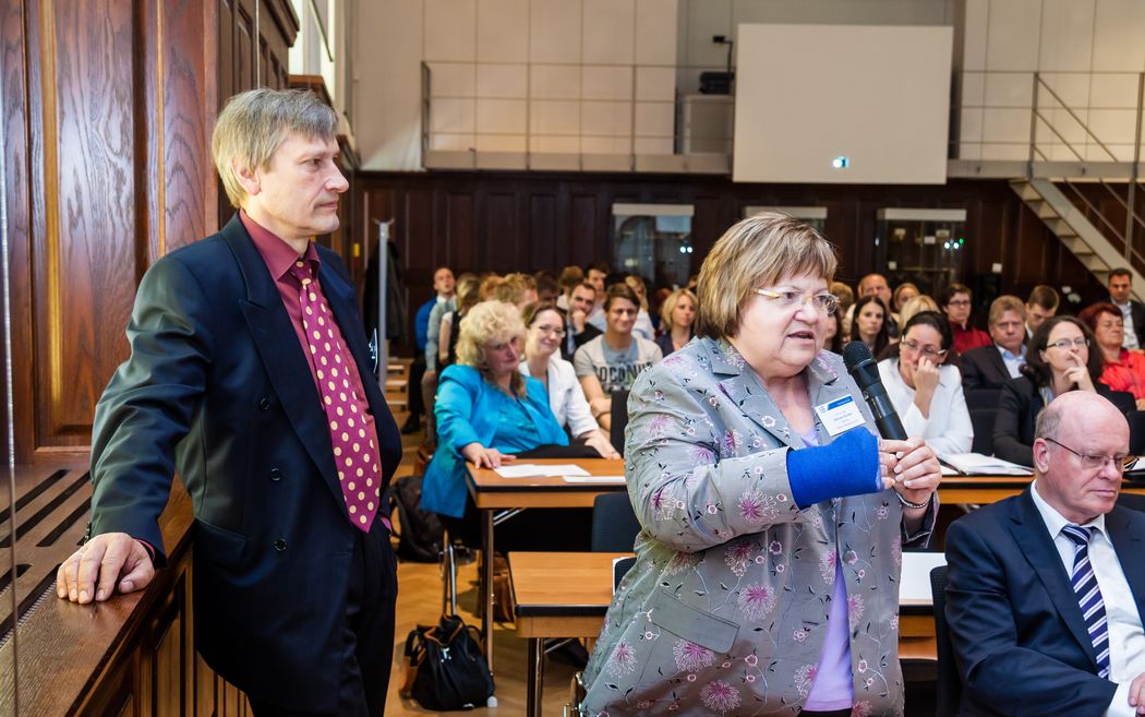 Foto: Zwei Teilnehmer stehend im Vordergrund. Dahinter weitere sitzende Teilnehmer.