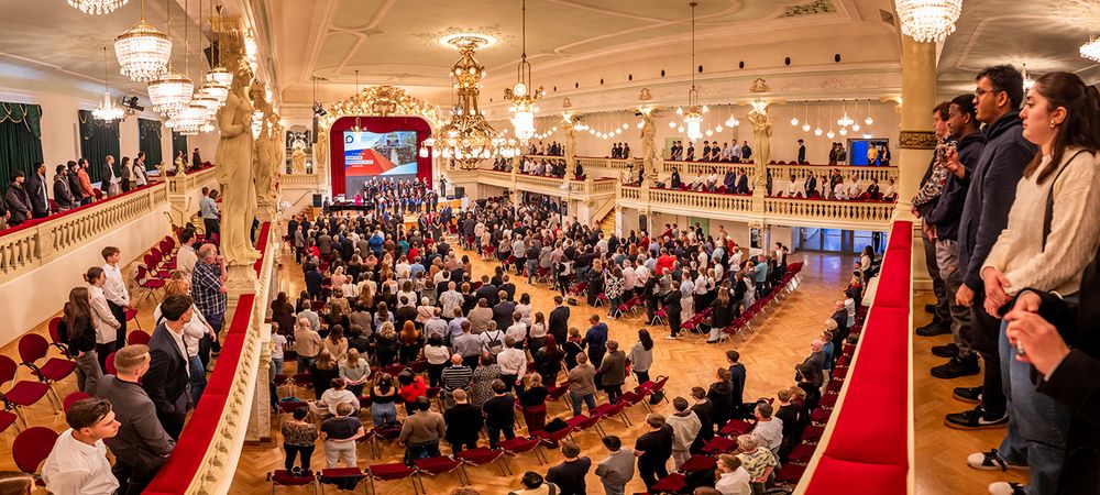 Blick in einen festlich beleuchteten Saal. Der Saal ist voll, das Publikum steht. 