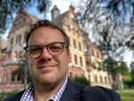 Foto: Im Vordergrund ist das Gesicht eines Mannes, im Hintergrund ein altes Gebäude.