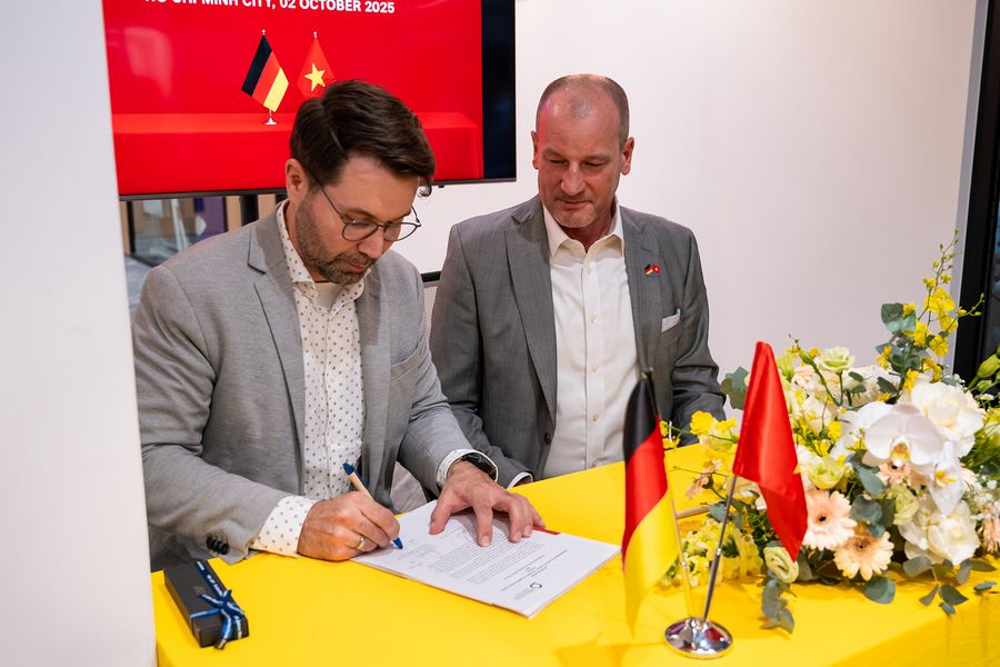 Zwei Personen in Anzügen sitzen an einem Tishc und unterzeichnen ein Dokument. Auf dem Tisch eine dutsche und eine vietnamesische Flagge. 