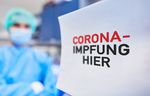 Foto: Vorn rechts ist ein Schild mit dem Text Corona-Impfung hier, im Hintergrund links steht eine Person in medizinischer Schutzkleidung und mit Mundschutz.