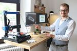 Foto: Ein junger Mann steht vor einem Schreibtisch. Auf dem sind ein 3D Drucker und zwei Monitore zu sehen.
