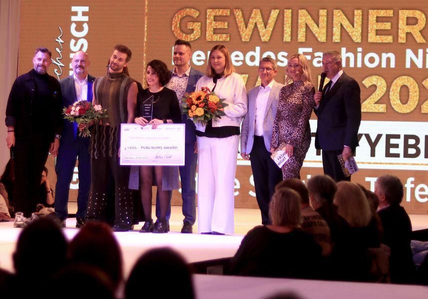 Eine Gruppe von Personen steht auf einer hell erleuchteten Bühne, im Hintergrund der Schwirftzug "Gewinner" 