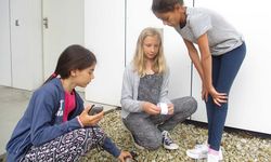 Foto: Drei Mädchen in einem Raum. Eine hält eine Stoppuhr, eine andere rollt Papier in ihren Händen auf und das dritte Mädchen schaut zu.
