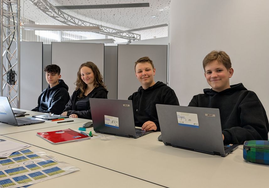 drei Schüler und eine Schülerin sitzen an ihrem Notebook und blicken in die Kamera