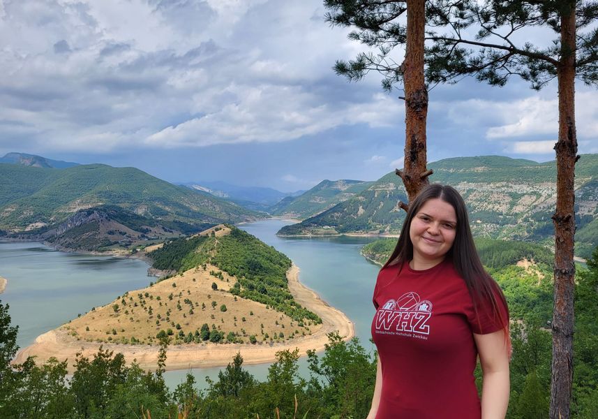Studierende im Hochschulshirt blickt in Kamera, im Hintergrund Flusslandschaft